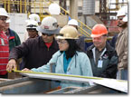 Mrs. Nazal, wife of former SUPSHIP accounting tech Bert Nazal, measures the first metal part cut for T-AKE 11.
